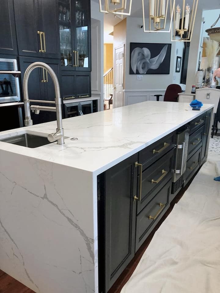 A kitchen island with a white marble-patterned countertop and sink, contrasted by dark cabinets with gold hardware.