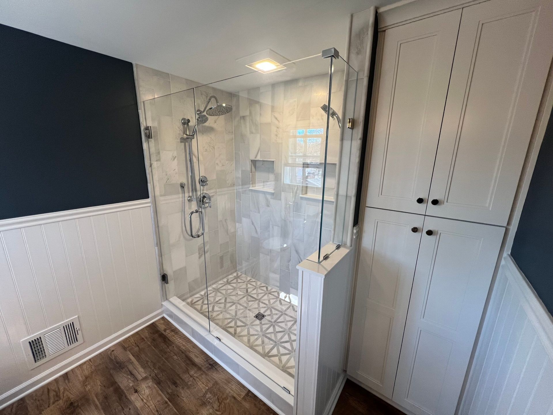 A modern walk-in shower with glass panels, patterned tile flooring, and light-colored cabinetry in a bathroom.