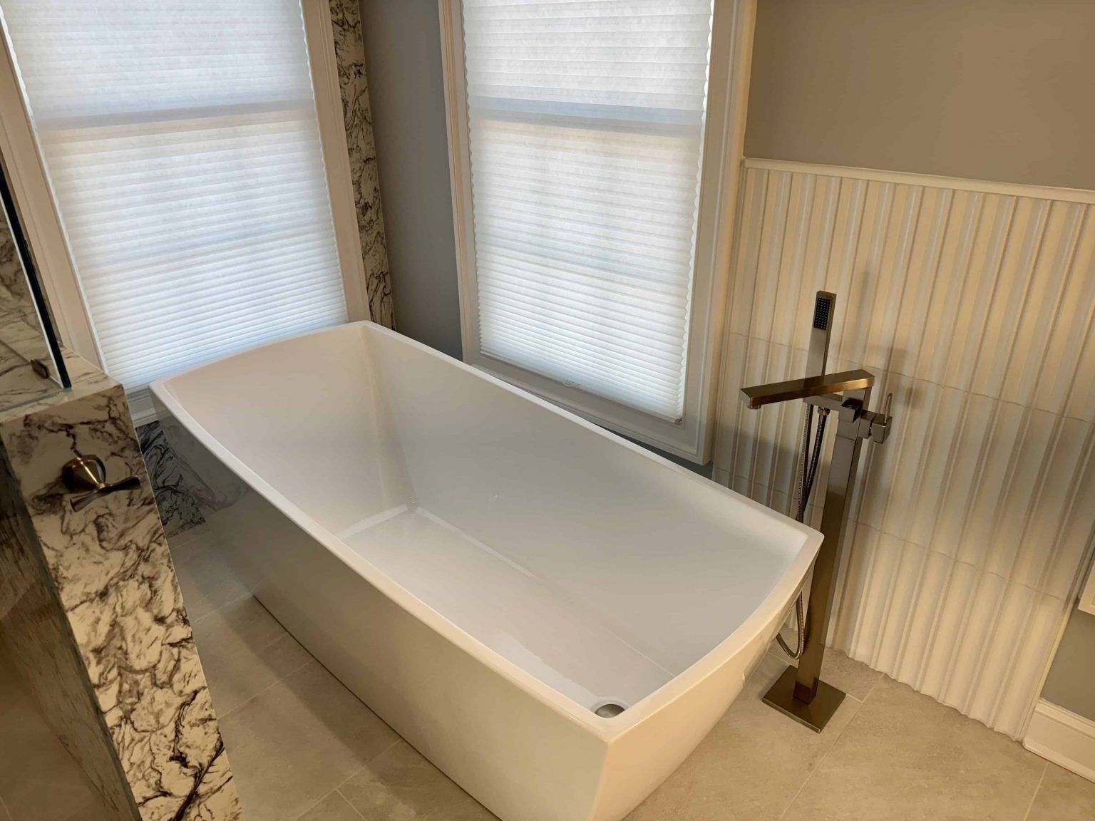 A white freestanding bathtub sits in a bathroom next to a tall brass floor-mounted faucet and windows with white blinds. A white freestanding bathtub sits in a bathroom next to a tall brass floor-mounted faucet and windows with white blinds.