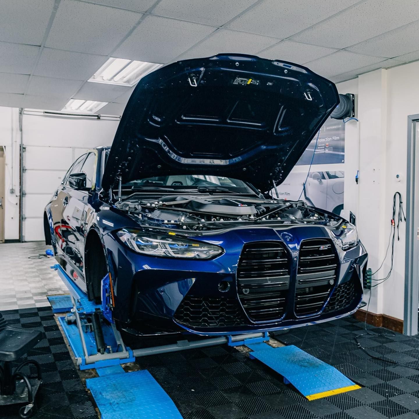 A blue car with the hood up is on a lift in a garage