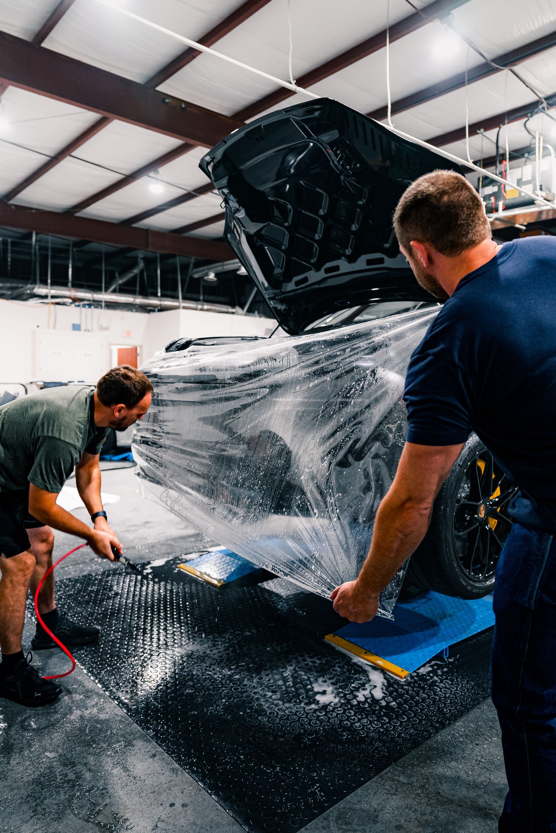 Two men are wrapping a car in plastic in a garage.