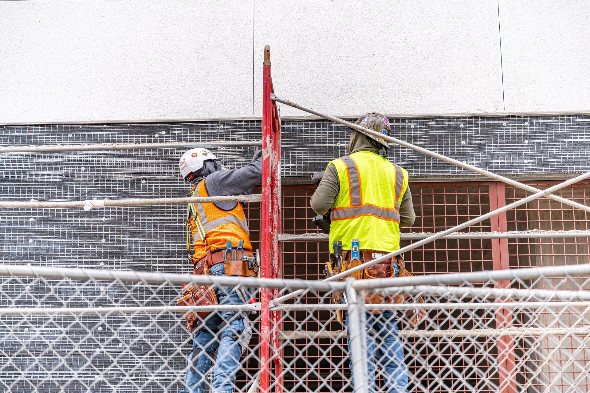 Two construction workers in safety vests on scaffolding behind a chain-link fence