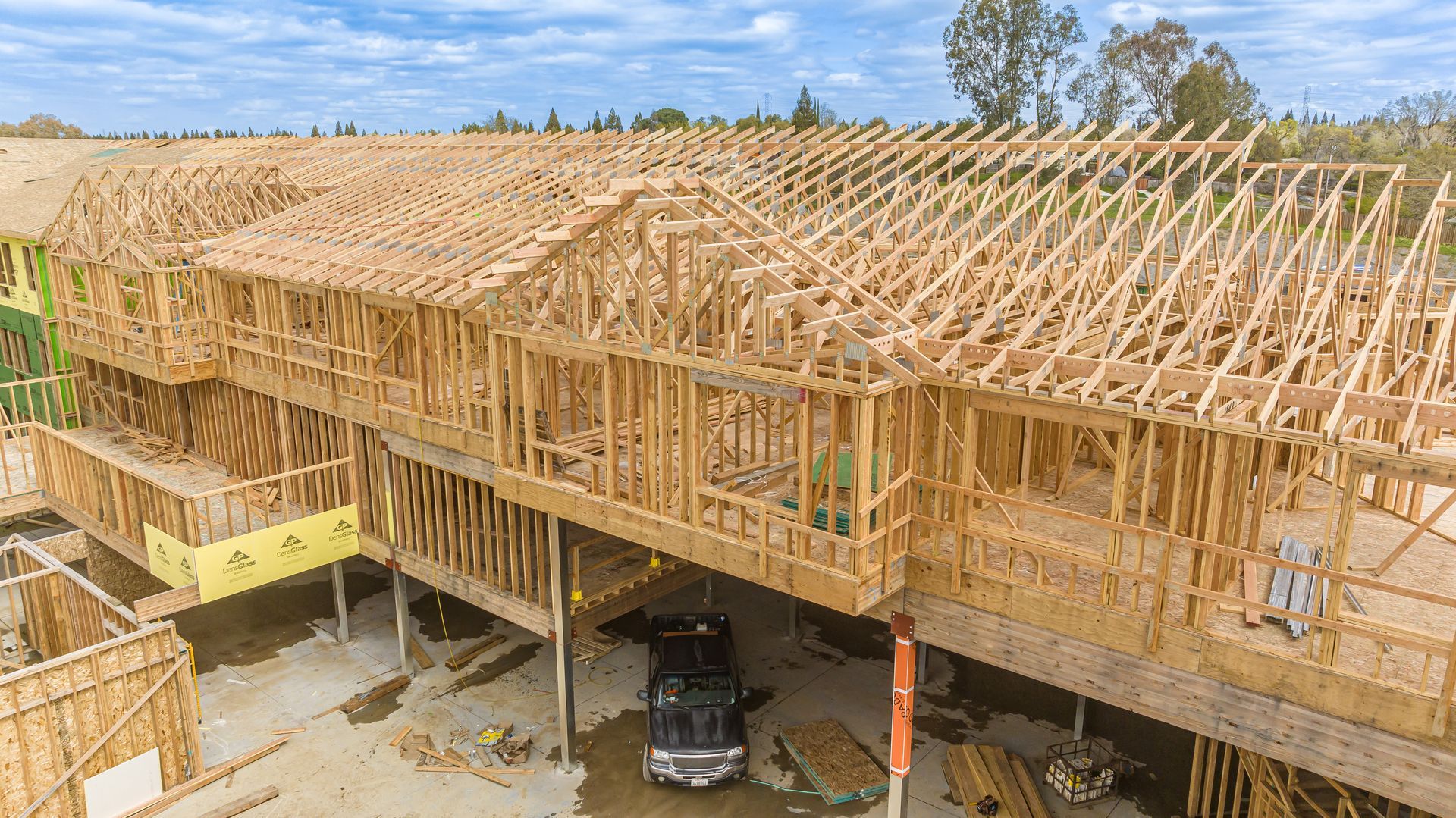 Large wooden house under construction with exposed roof trusses and scaffolding on a sunny day