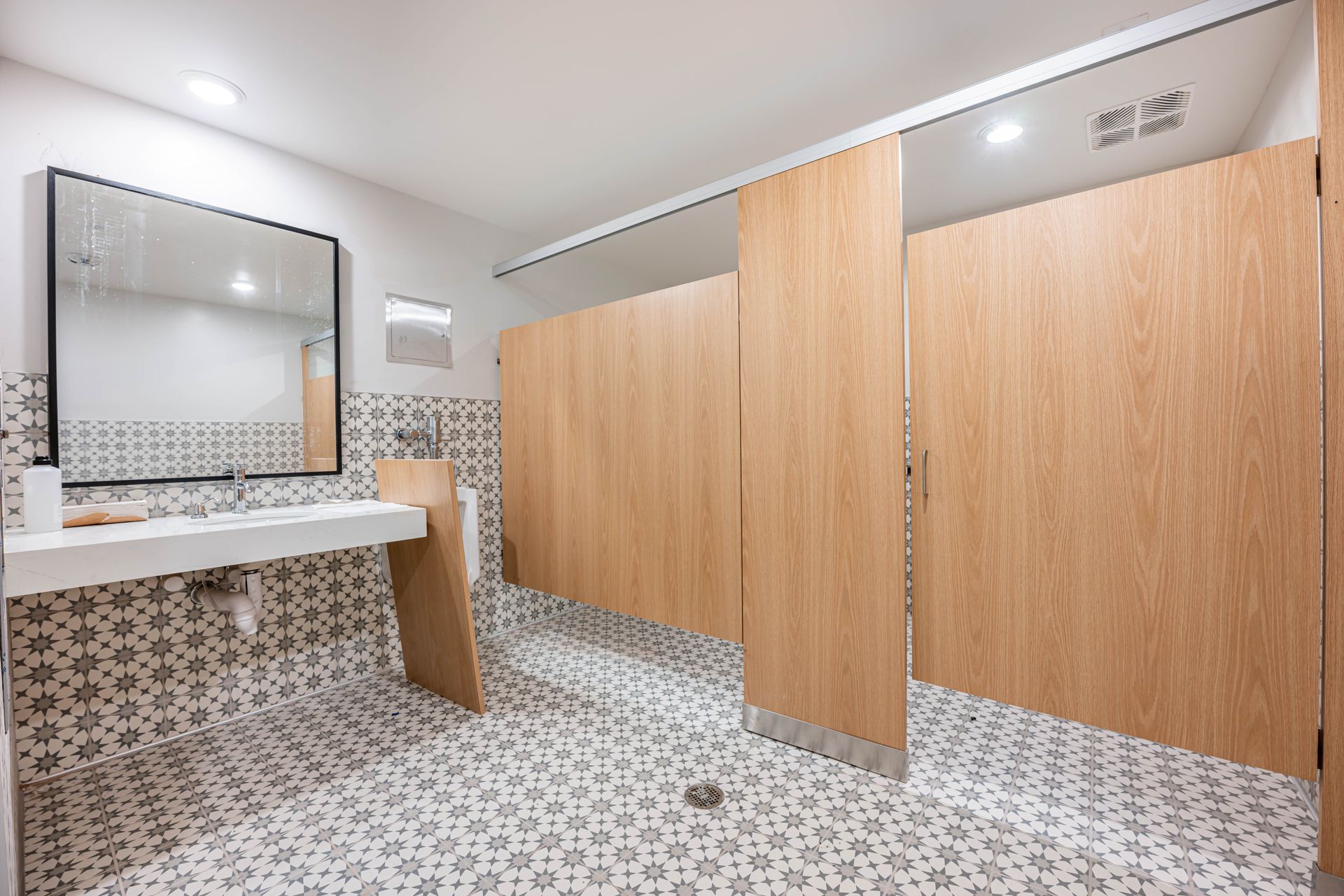 Modern restroom with patterned tile, a white sink counter, mirror, and wood stall doors