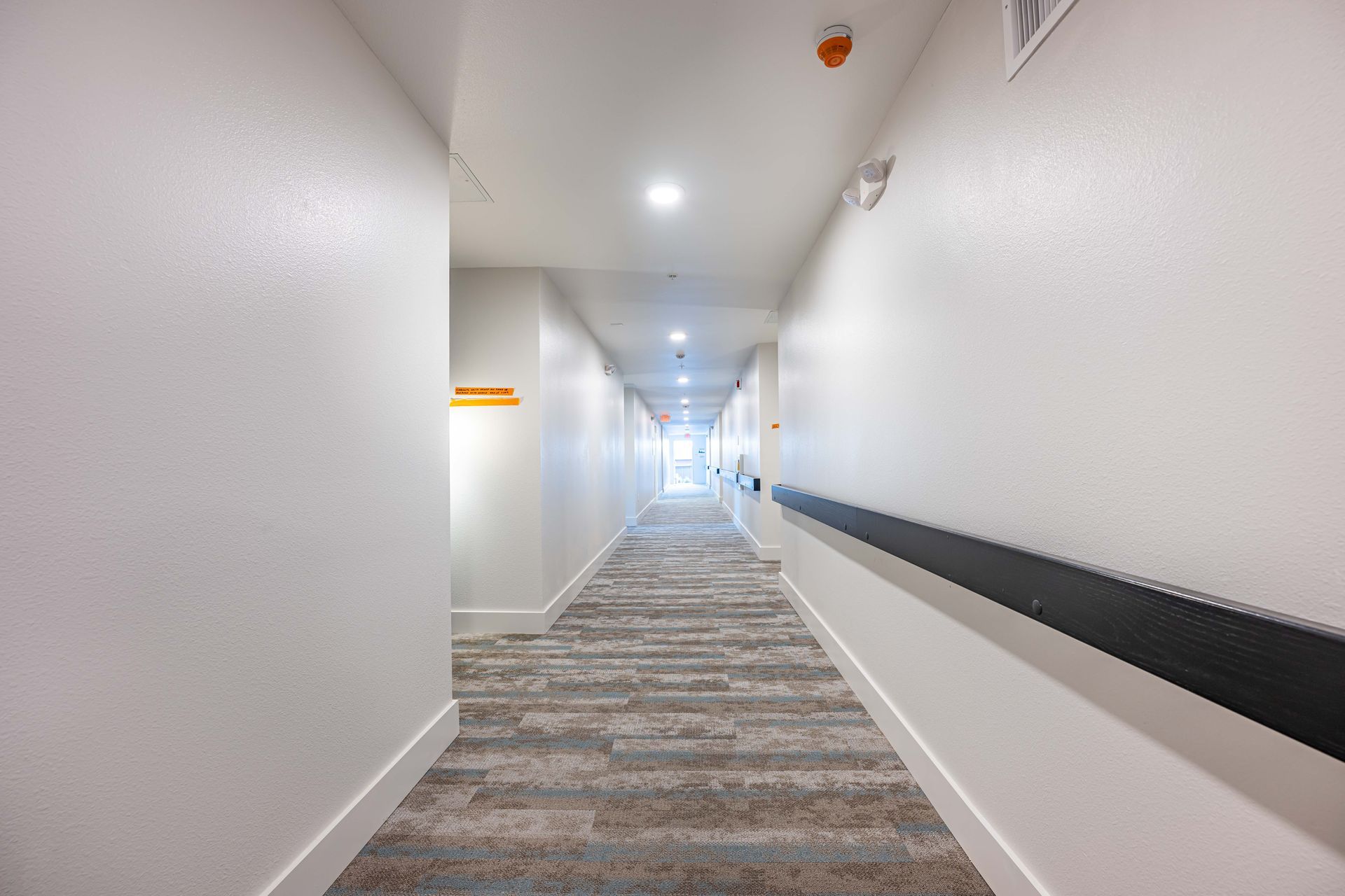 Bright hotel hallway with white walls, patterned carpet, and recessed ceiling lights, receding into the distance