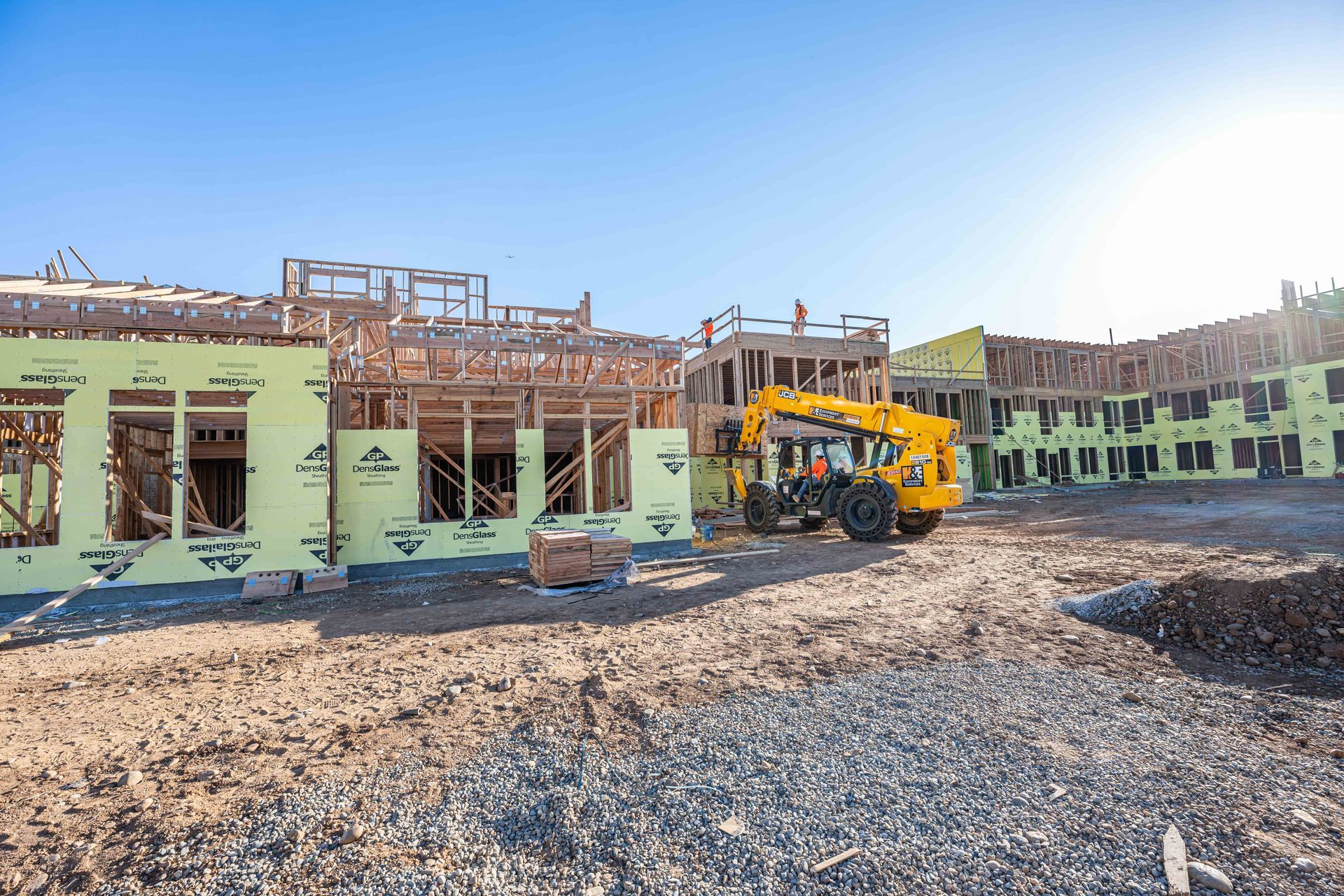 Construction site with a yellow excavator beside a partially built green building under a clear blue sky