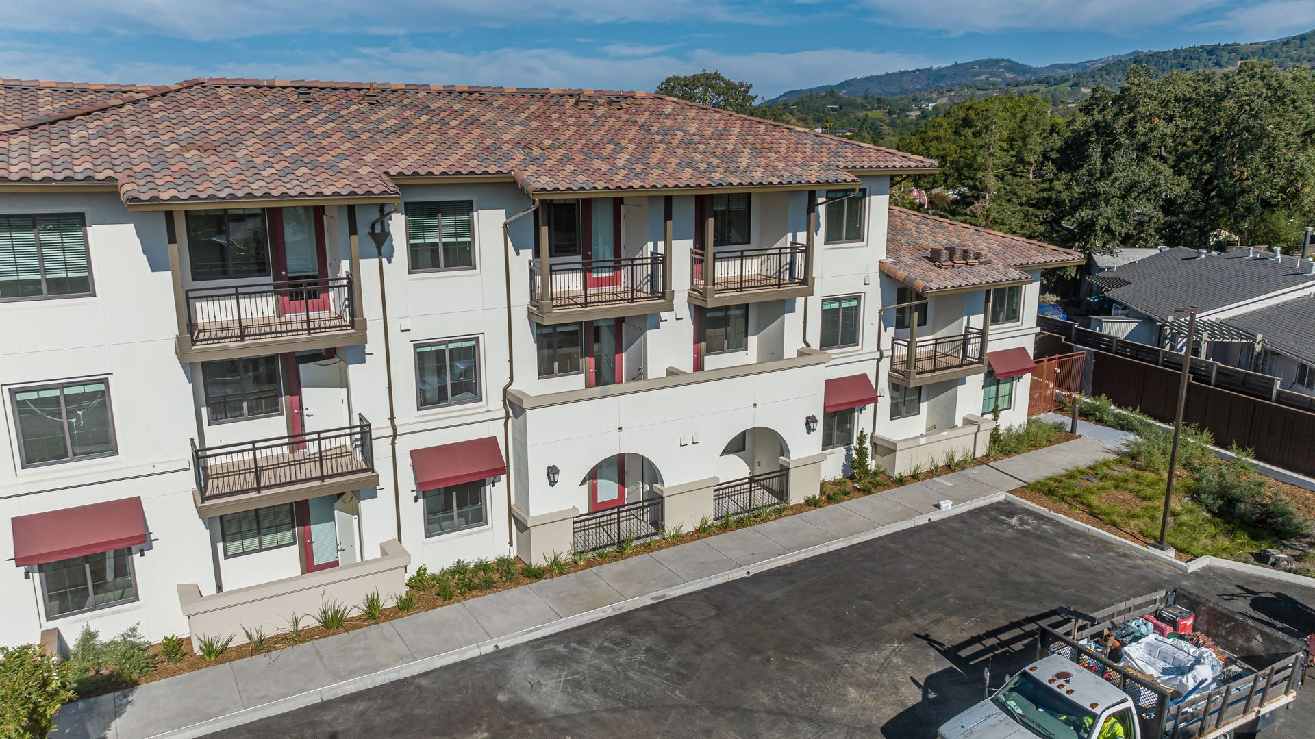 Three-story apartment building with red-tile roof, balconies, and parking lot in front.