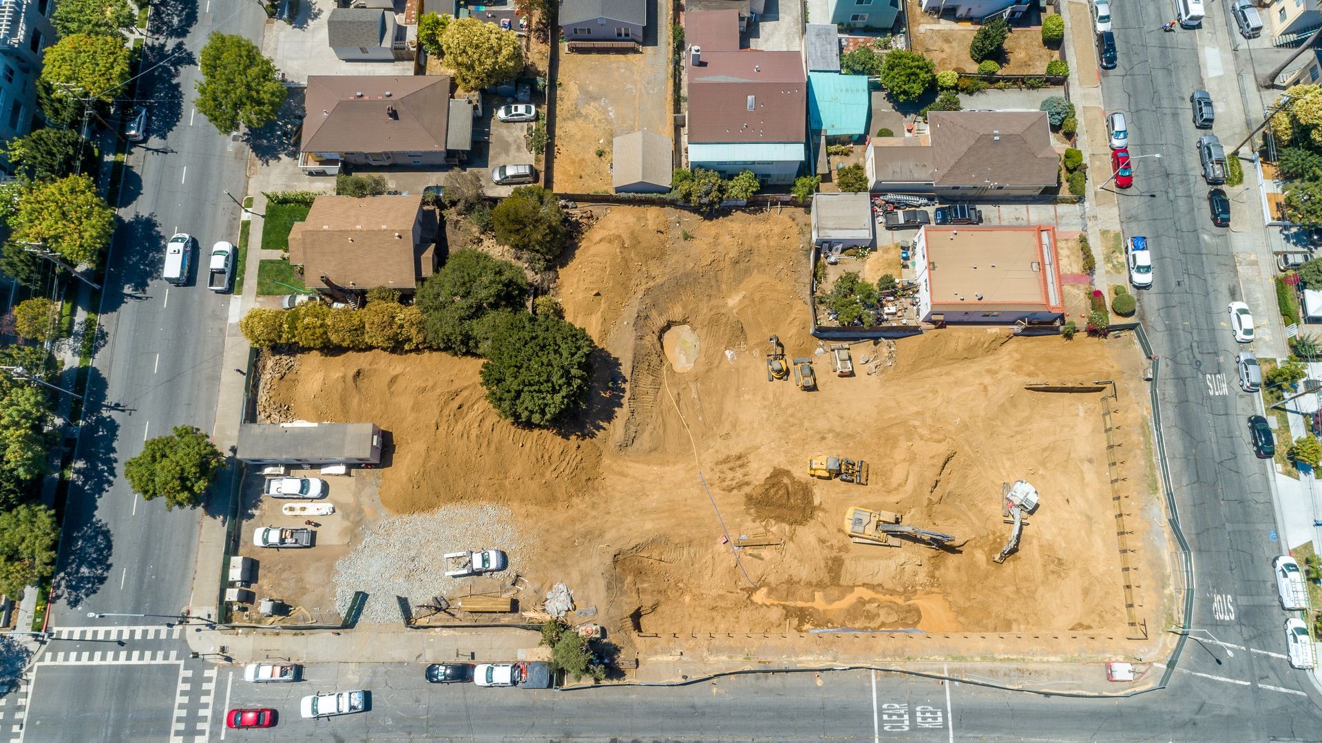 Aerial view of a vacant lot under construction beside houses and a street with cars.