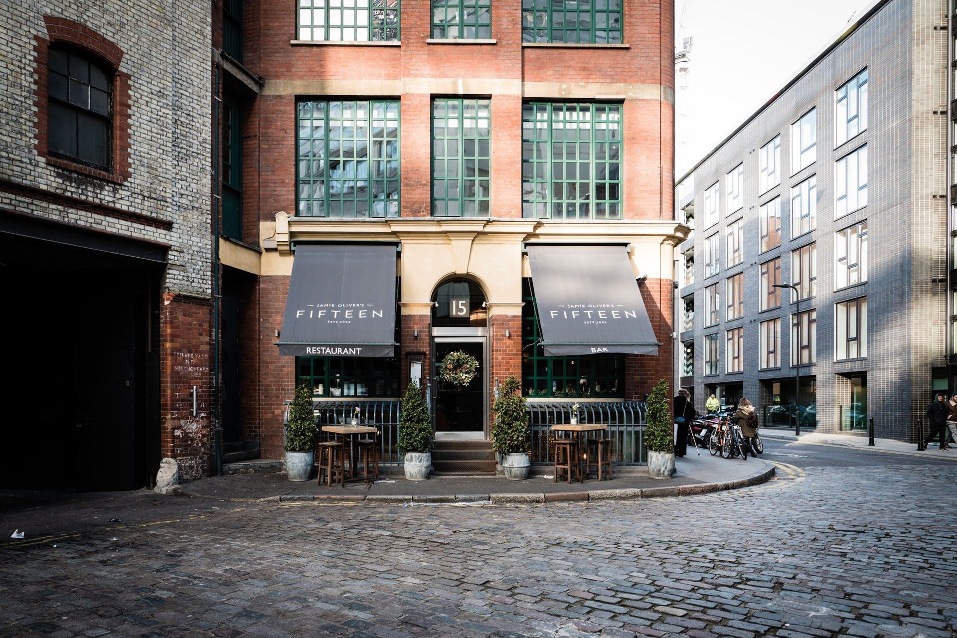 Cobblestone street with a red-brick café storefront and outdoor tables on a rainy city corner