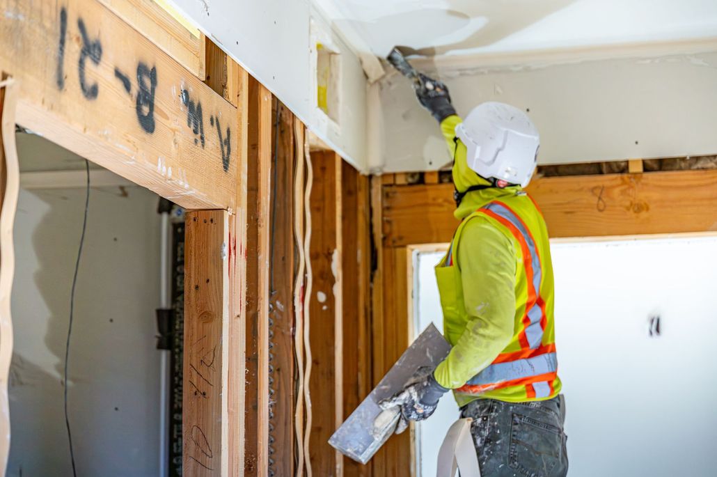 Construction worker wearing hard hat and safety vest applies material to a ceiling inside a house under renovation.