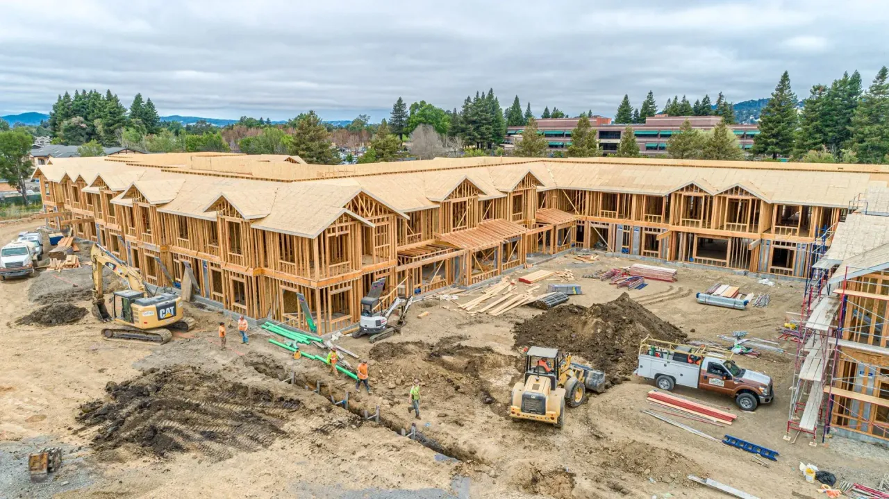Aerial view of a large multi-unit residential building under construction with heavy machinery on a dirt job site.