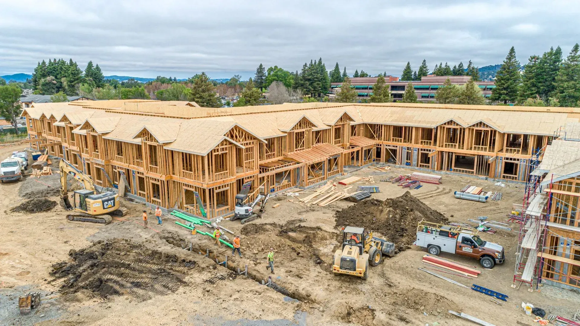 Aerial view of a large multi-unit residential building under construction with heavy machinery on a dirt job site.