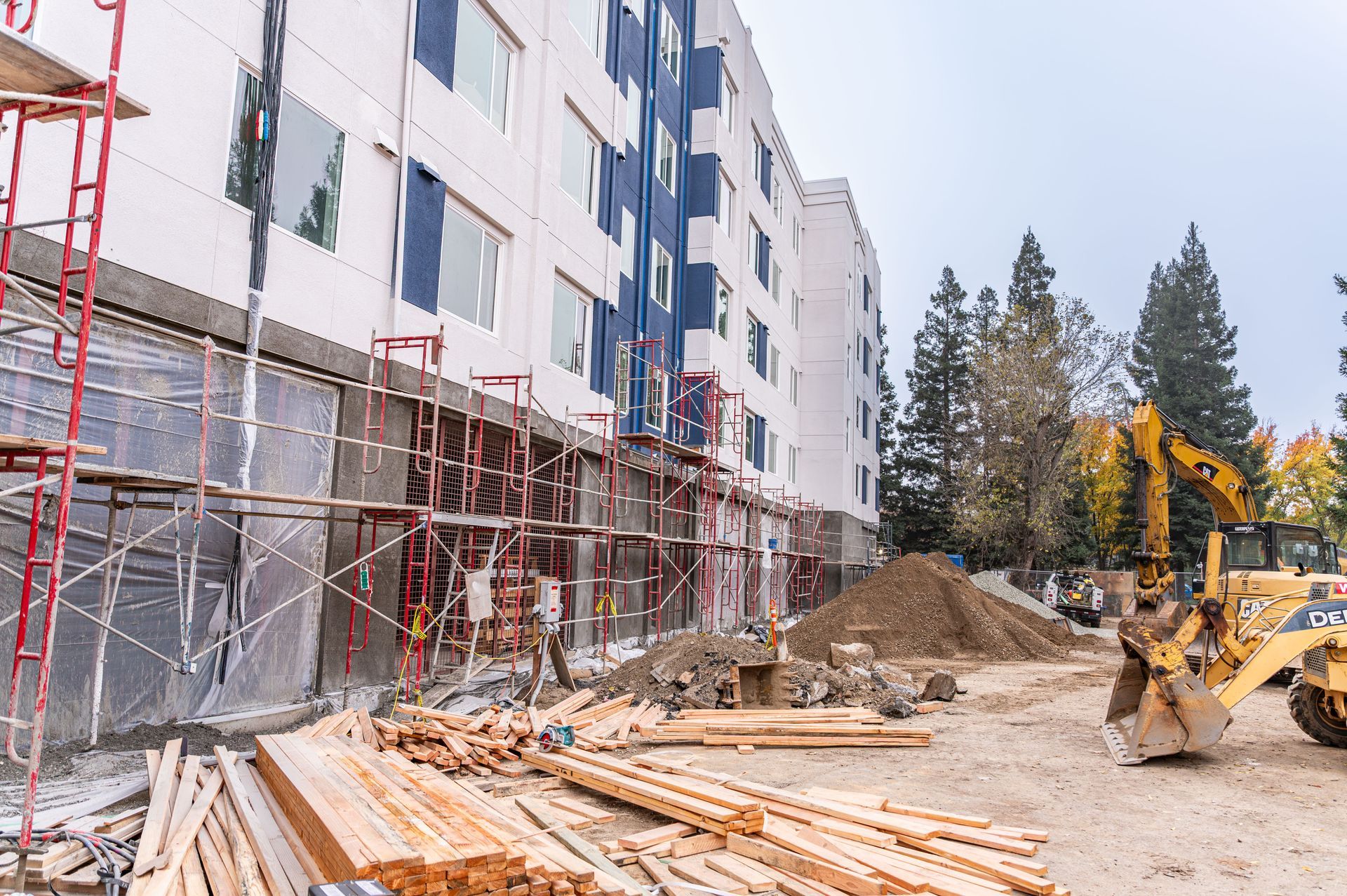 Apartment building under construction with scaffolding, lumber piles, and an excavator on a dirt site.
