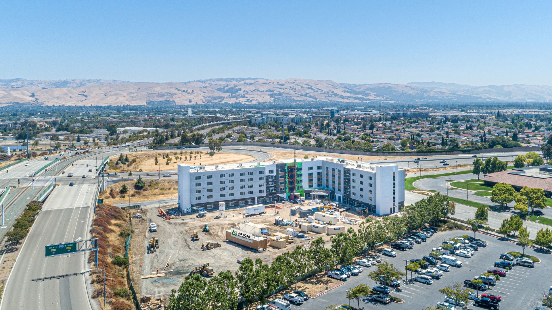 Aerial view of a modern building complex under construction beside a parking lot and road in a desert city.