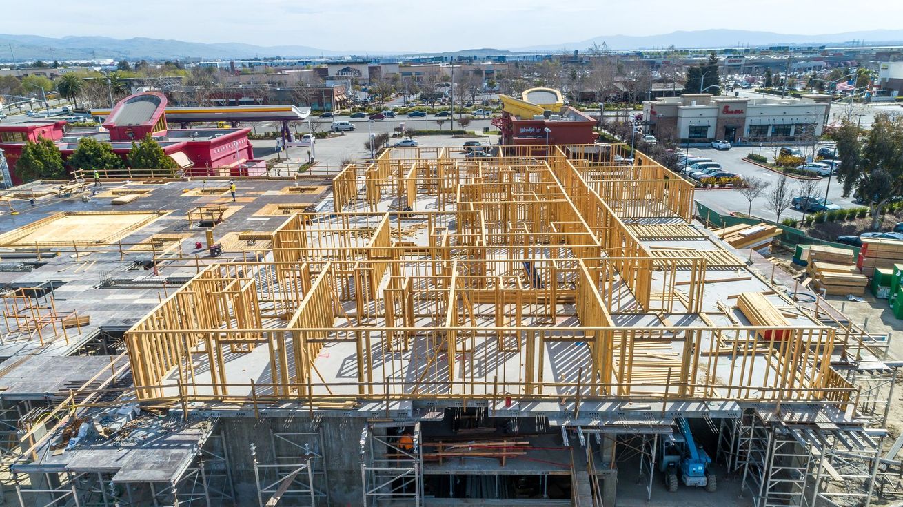 Aerial view of a building under construction with wooden roof framing and surrounding urban landscape