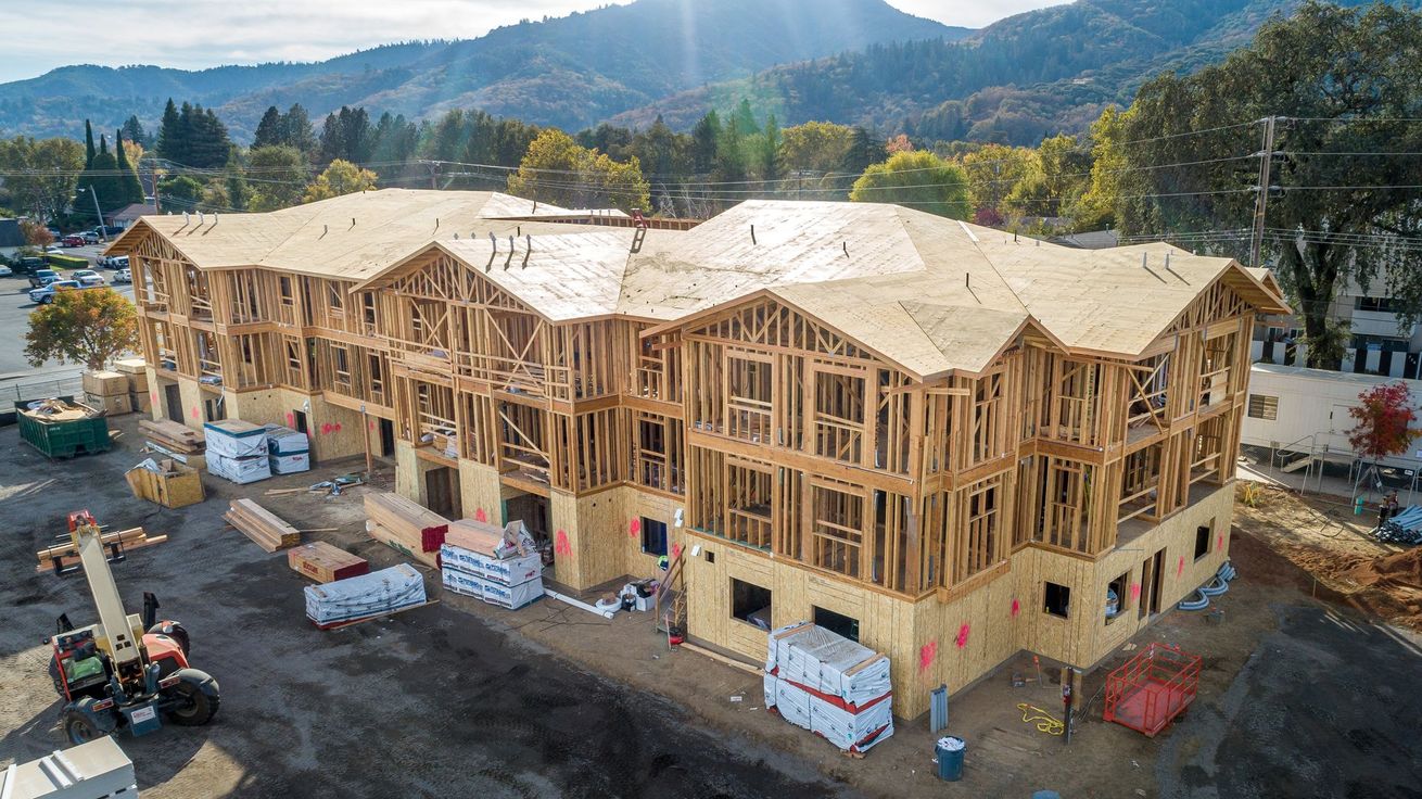 Wood-framed house under construction at a hillside site with mountains in the background