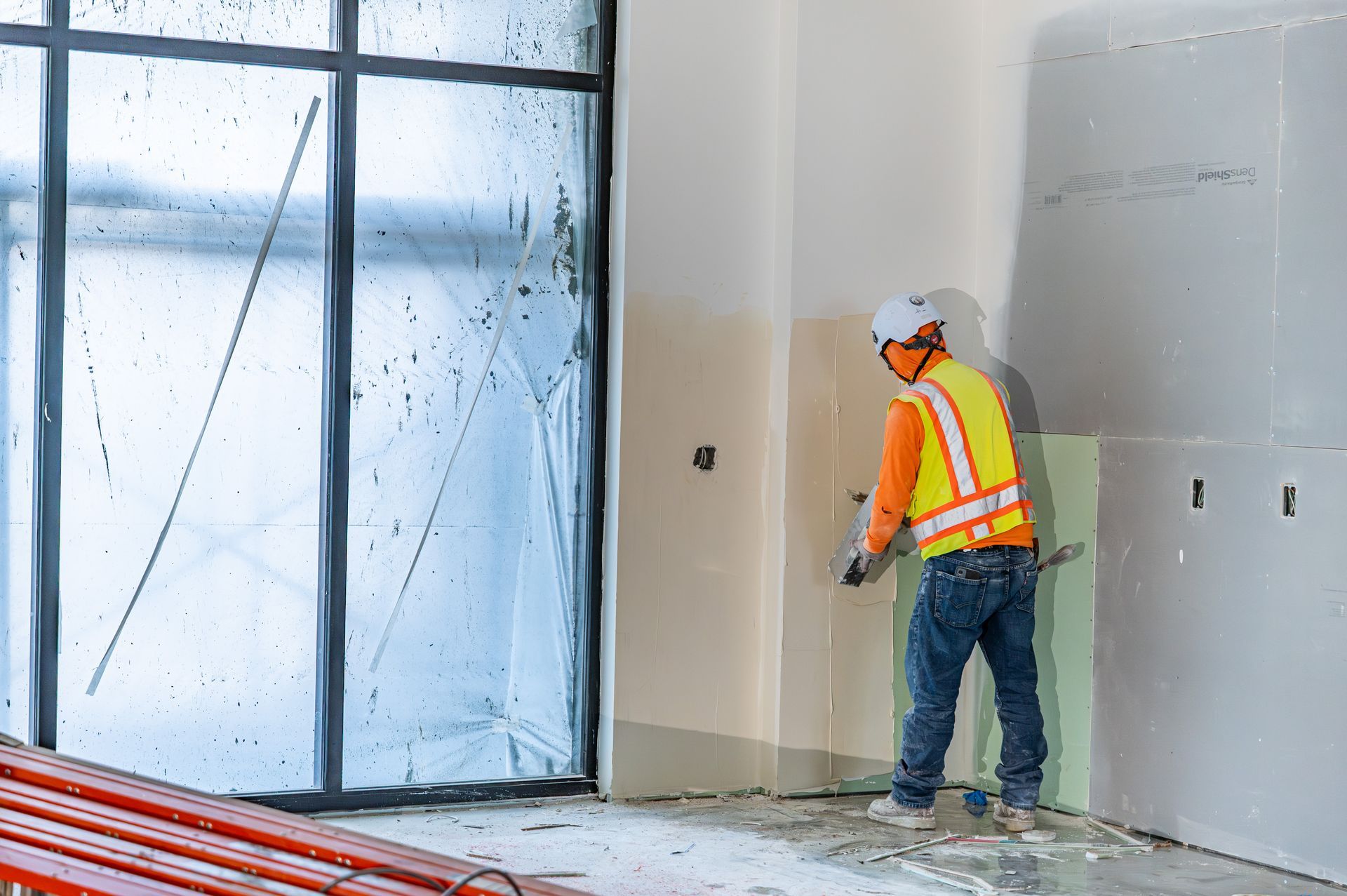 Construction worker in orange safety vest standing by a wall near a large window at an unfinished site