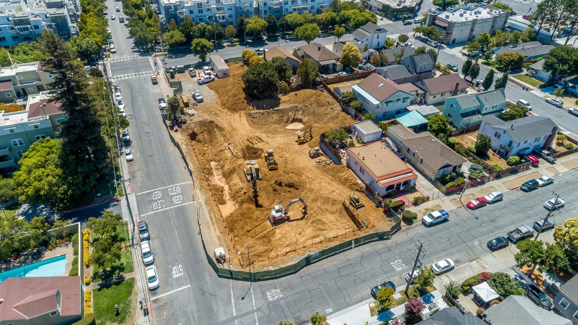 Aerial view of a vacant dirt lot beside streets and homes, with cars and trees around it.