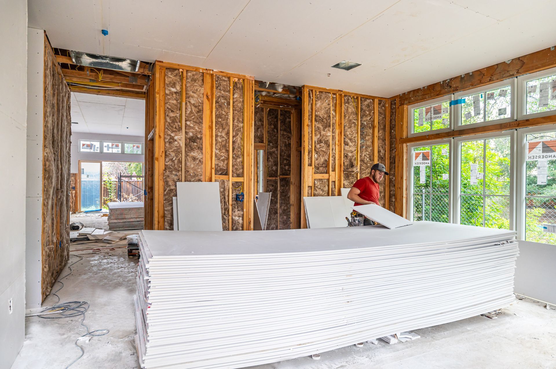 Partially renovated room with exposed framing, white counter, and a worker in a red shirt near large windows