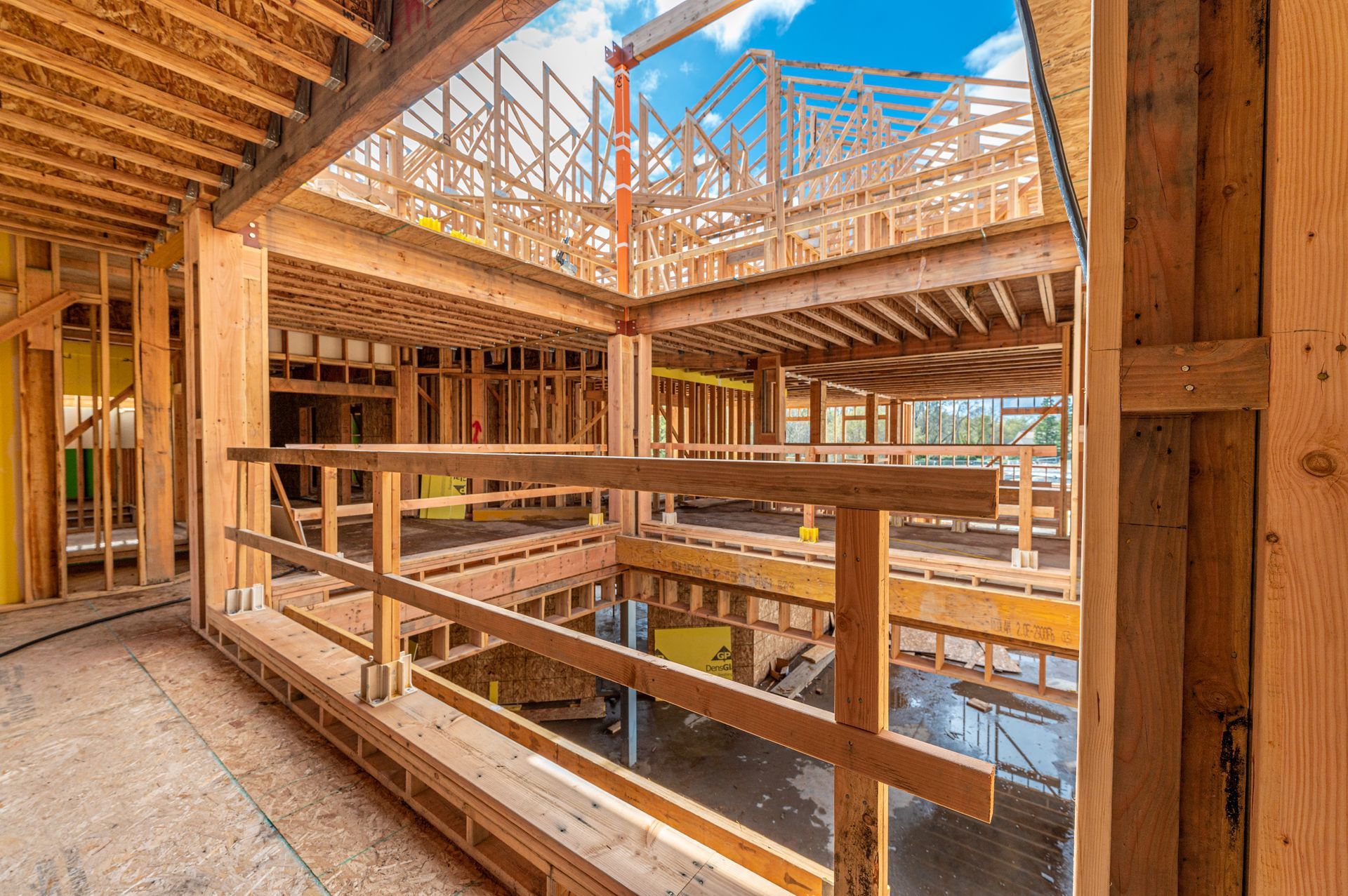 Interior of a wood-framed building under construction, with exposed beams, railings, and a skylight open to blue sky