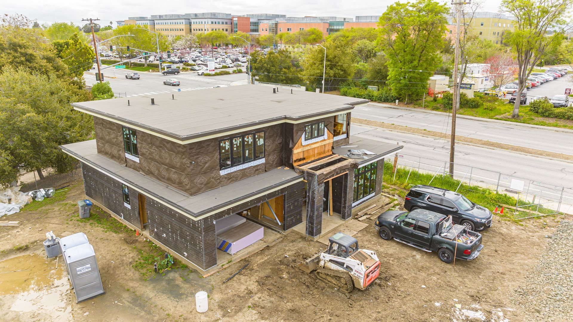 A modern two-story house under construction beside a road, with cars parked on site and trees around it.