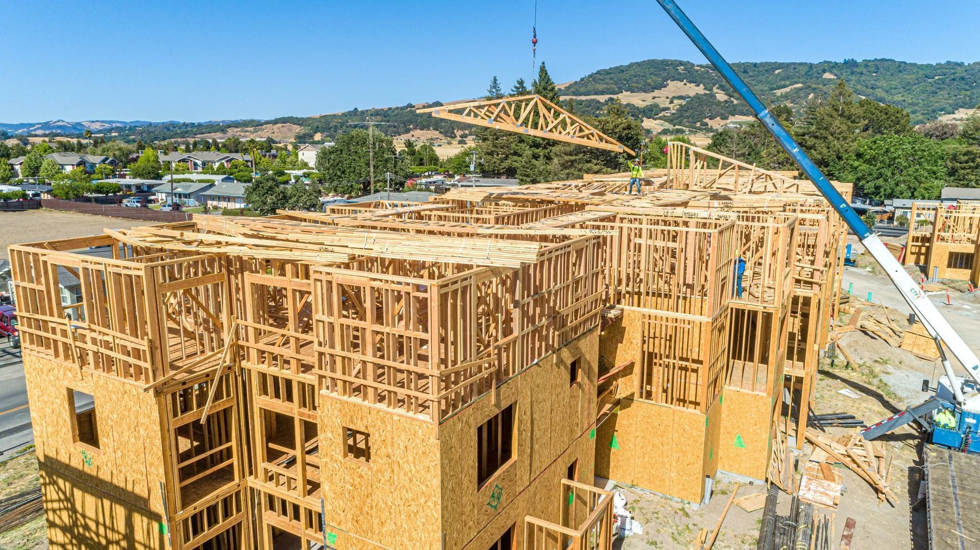 A construction crane hoists a wooden roof truss over a multi-story apartment building frame under a clear blue sky.