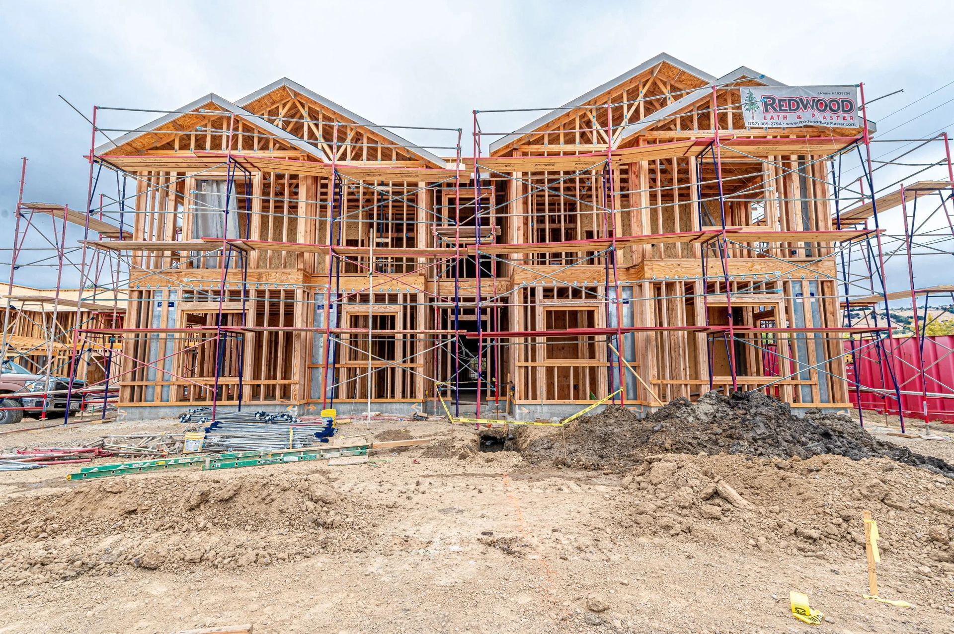 A wooden building frame under construction on a dirt lot, surrounded by scaffolding against a cloudy sky.