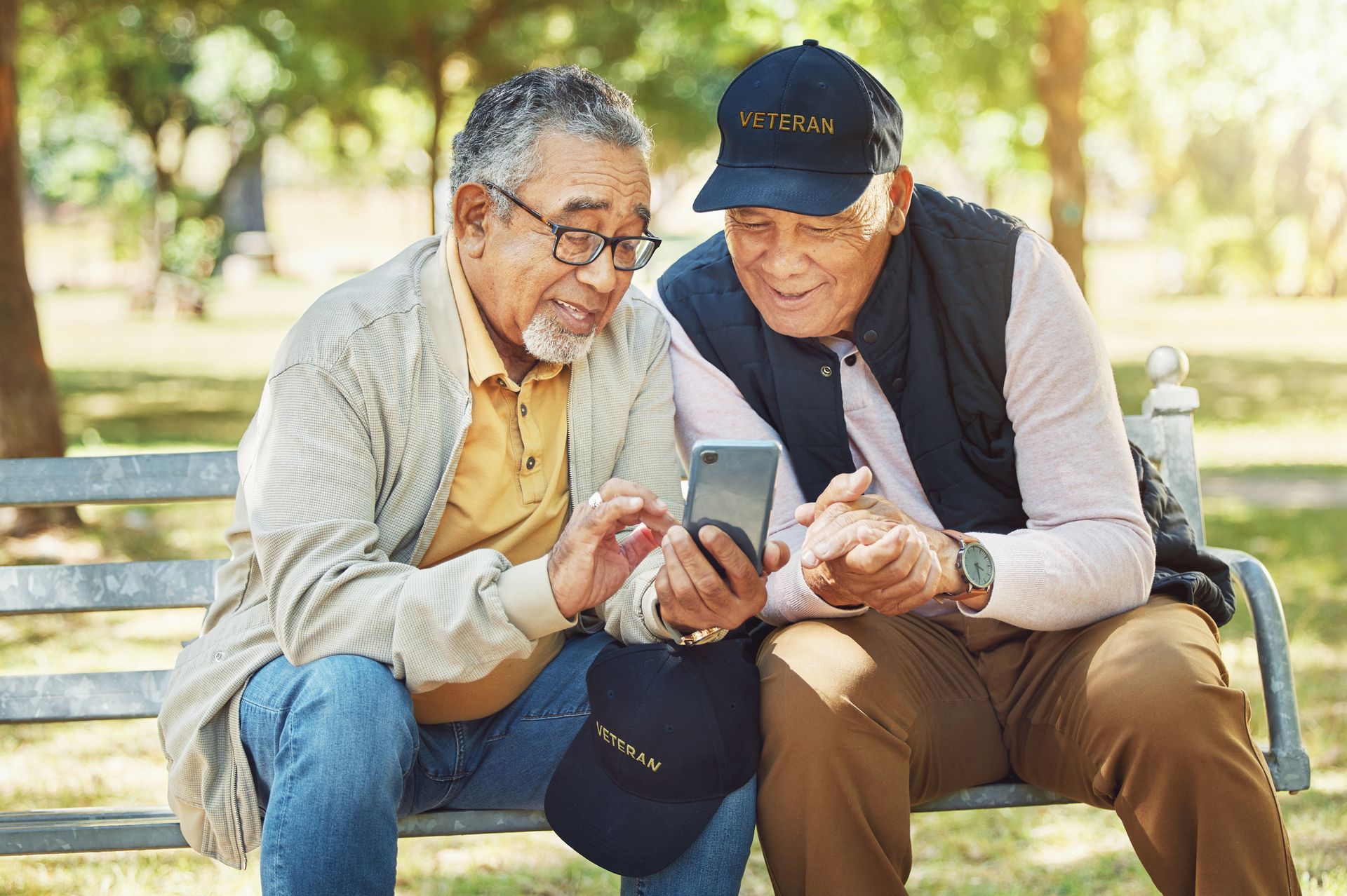 Two older men are sitting on a park bench looking at a cell phone.