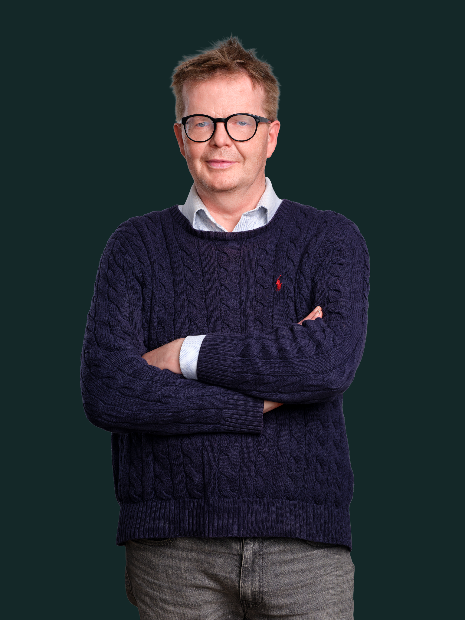 Andlitsmynd af manni í dökkbláum peysu og með gleraugu, standandi með krosslagða hendur á dökkgrænum bakgrunni