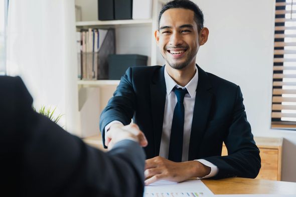 Man in suit shaking hands, smiling at a desk in an office.