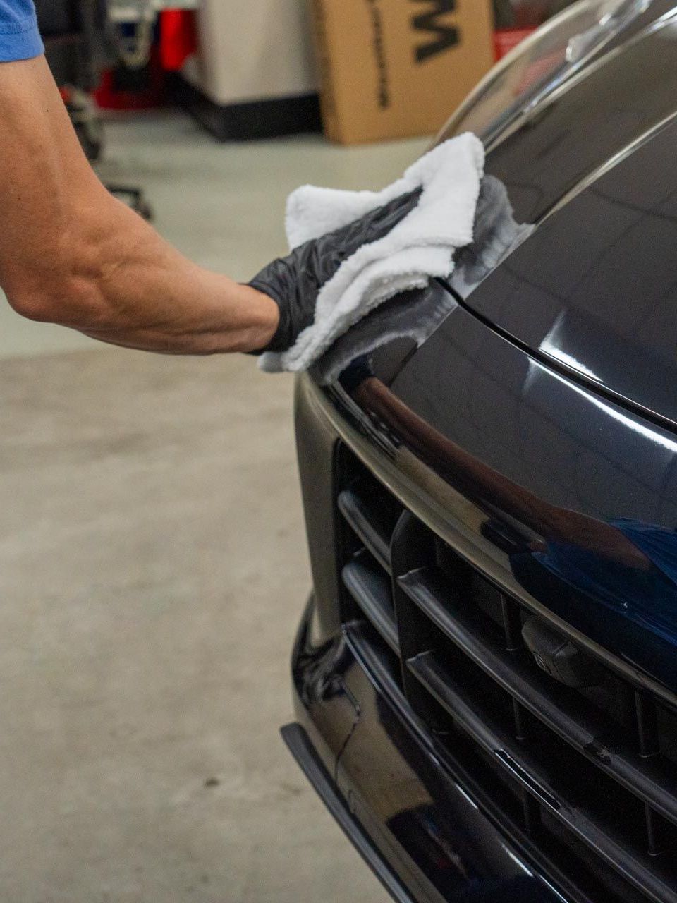 Gloved hand wiping a dark car bumper with a white cloth in a garage setting.