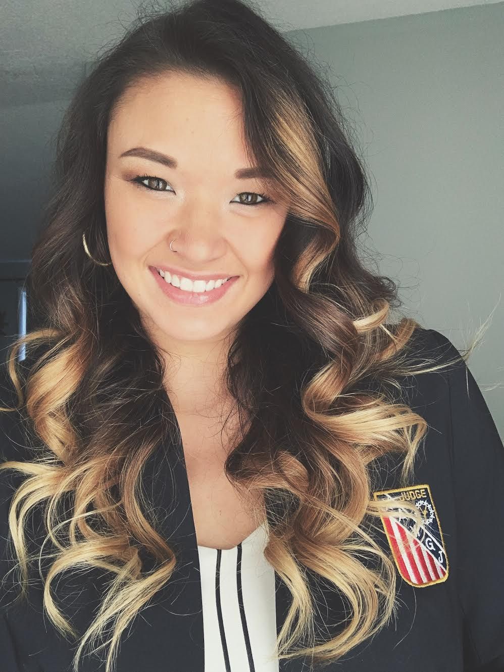 Smiling person with long wavy hair wearing a black top with a patriotic pin, indoors.