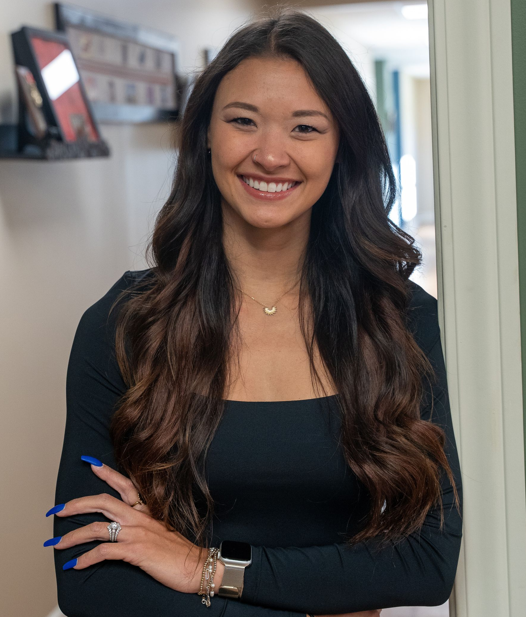 Smiling woman standing in a doorway, wearing a black top and long wavy hair, with a blurred office background