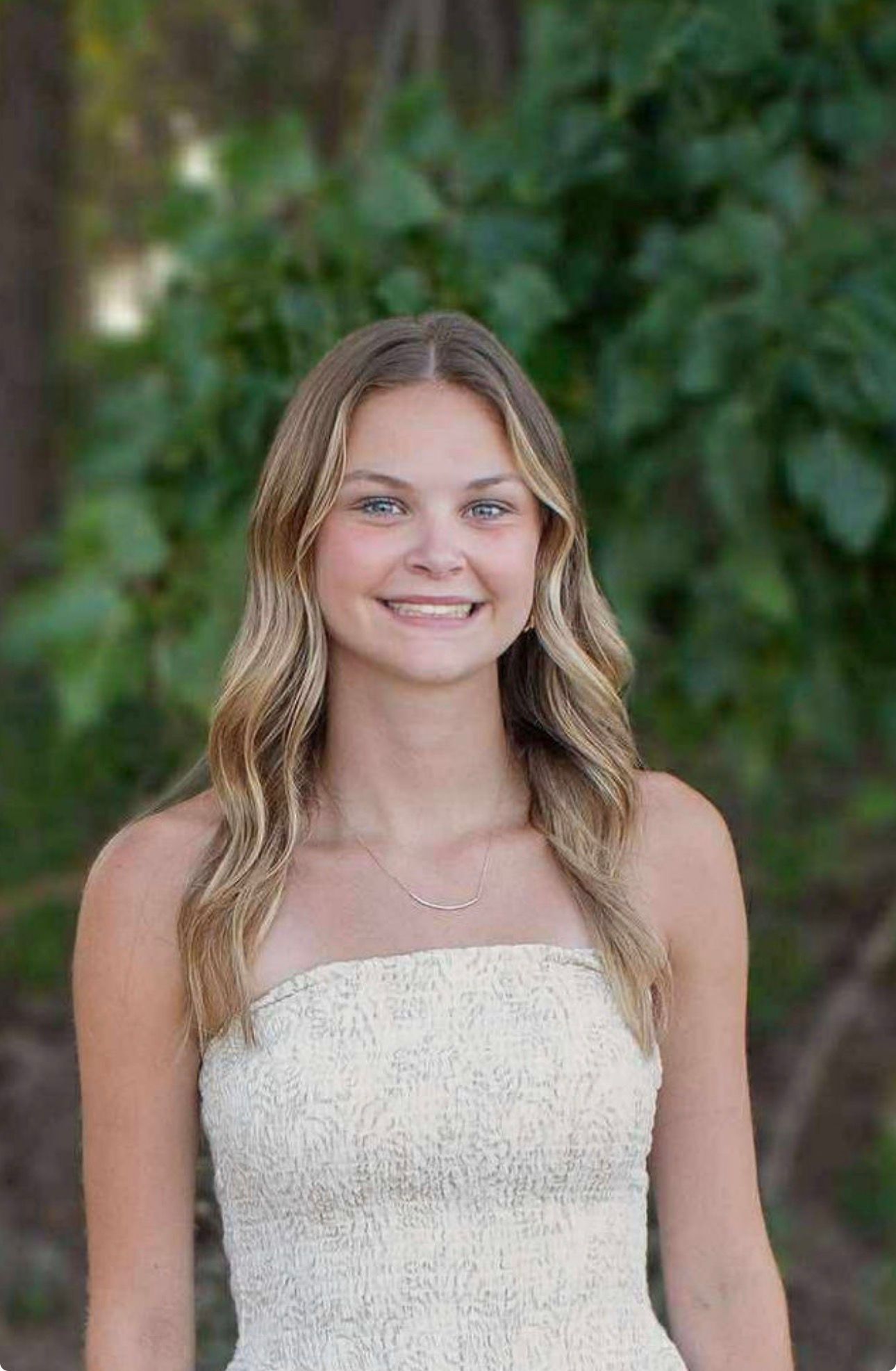 Smiling person in a white strapless dress standing outdoors against a leafy green background