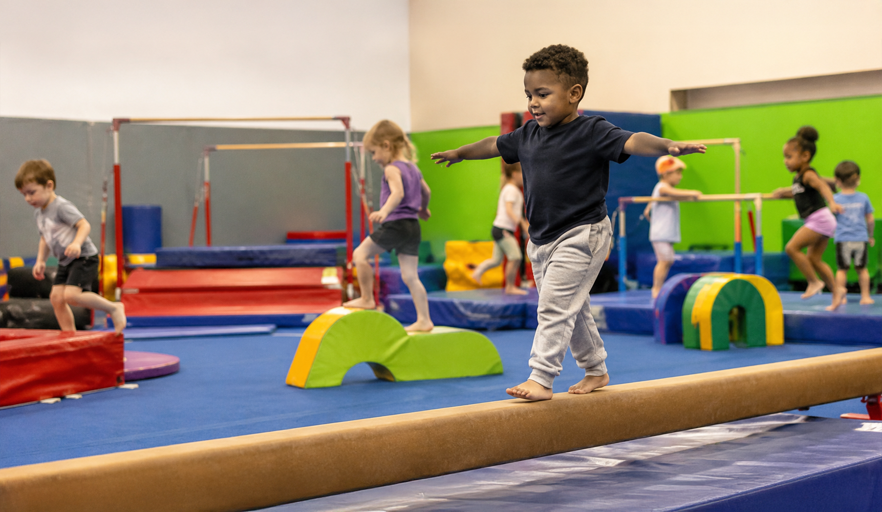 Children balancing on gym equipment in an indoor play area, with colorful mats and obstacles.