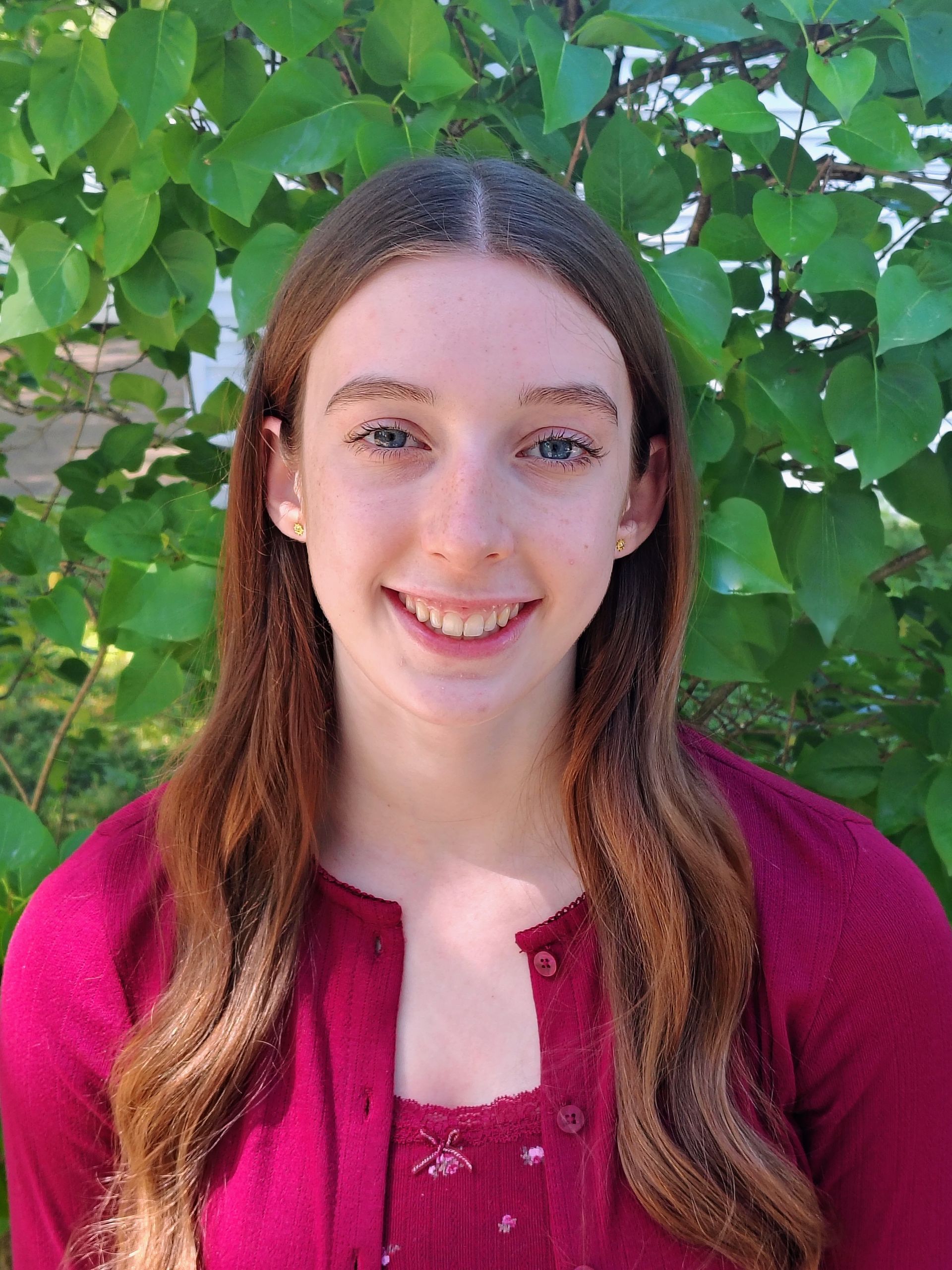 Smiling person with long brown hair in a magenta top standing in front of green leaves