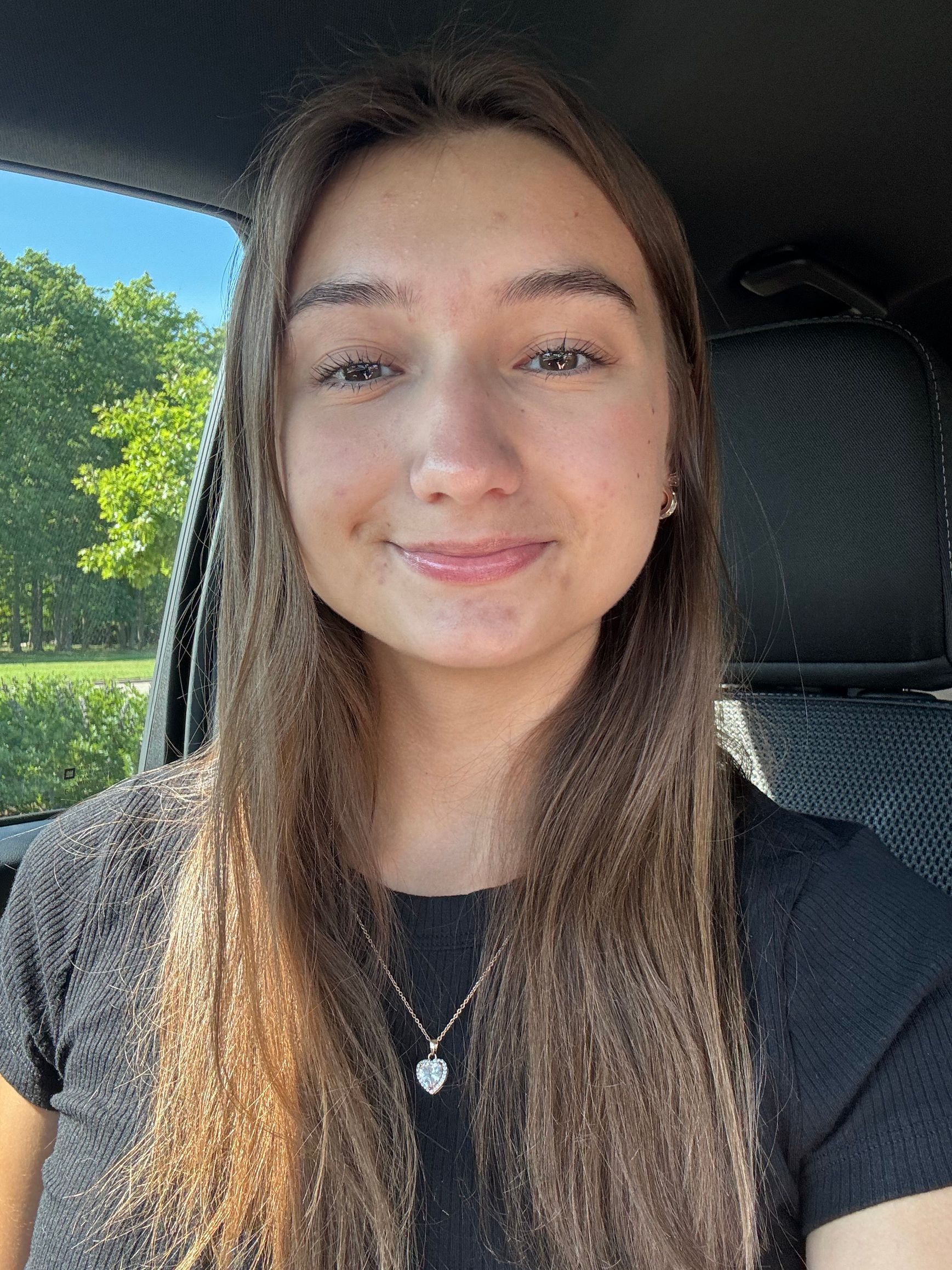 Smiling person with long hair sitting in a car, wearing a black shirt and necklace.