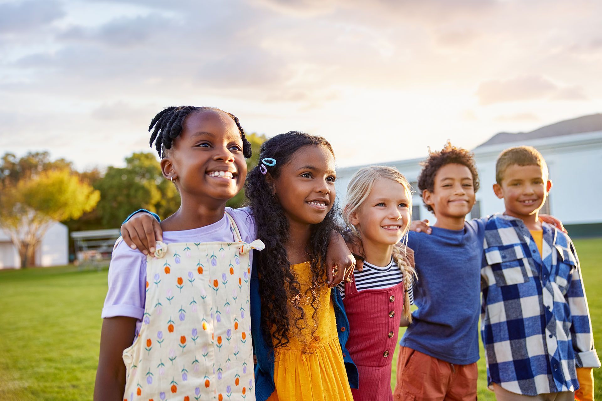 Five smiling children standing arm in arm outdoors at sunset