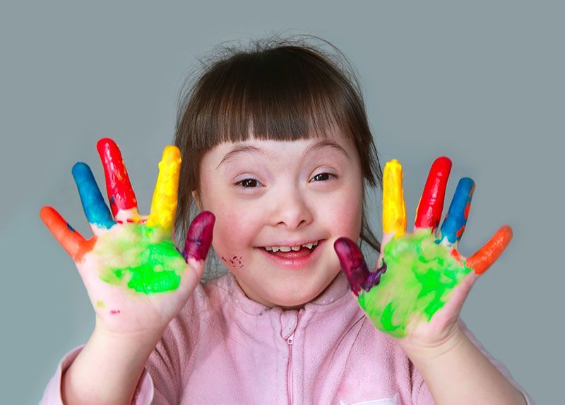 Smiling child with colorful paint on both hands, raising them against a gray background