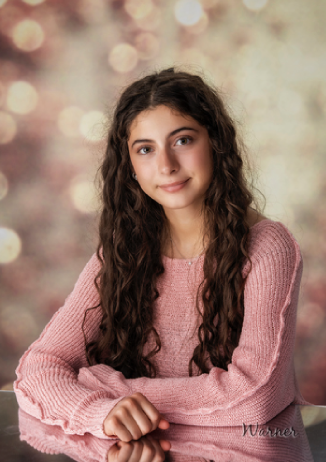Young person with long curly hair in a pink sweater, seated against a soft bokeh background, smiling gently