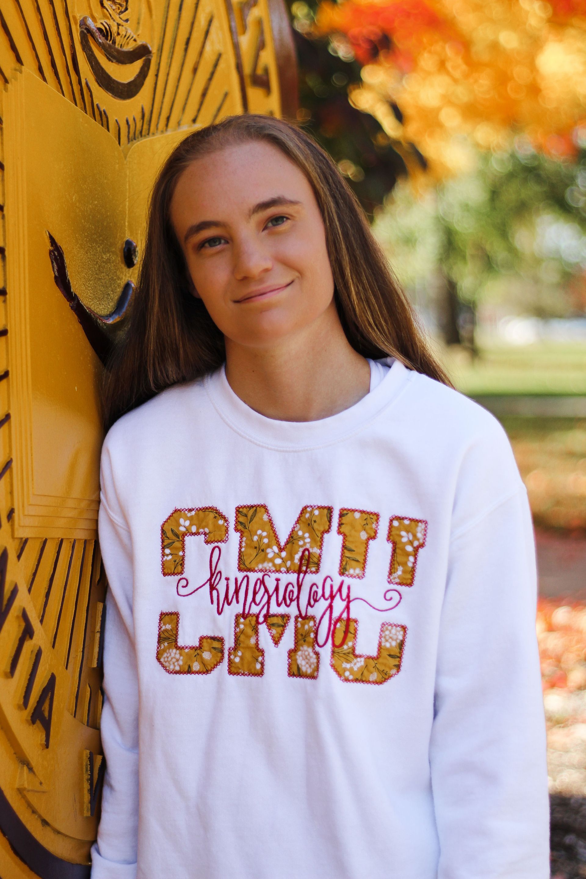 Smiling person in a white sweatshirt leaning against a yellow graffiti-covered wall outdoors.