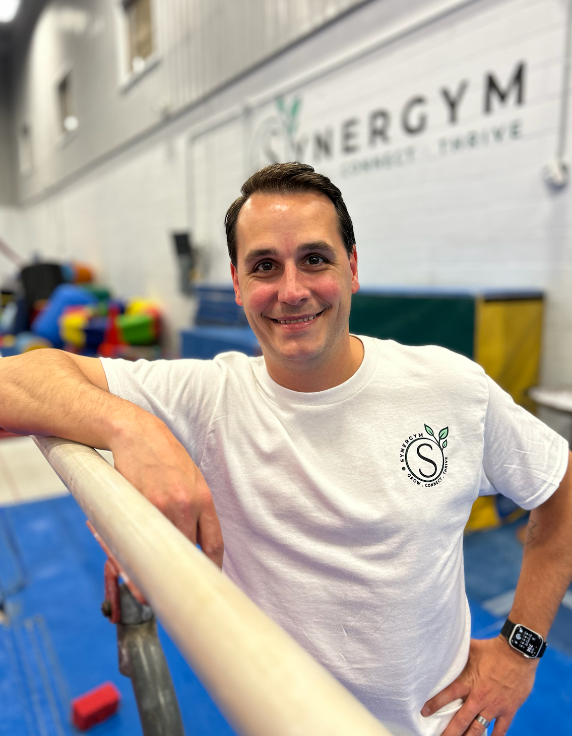 Man leaning on a gym railing, smiling, with an ENERGYM sign and colorful exercise equipment behind him