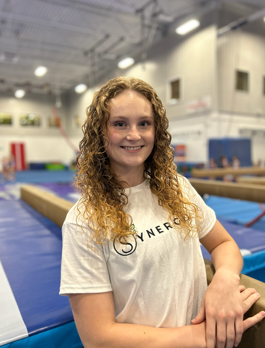 Smiling woman in a white T-shirt standing in a gymnastics gym with blue mats and equipment.