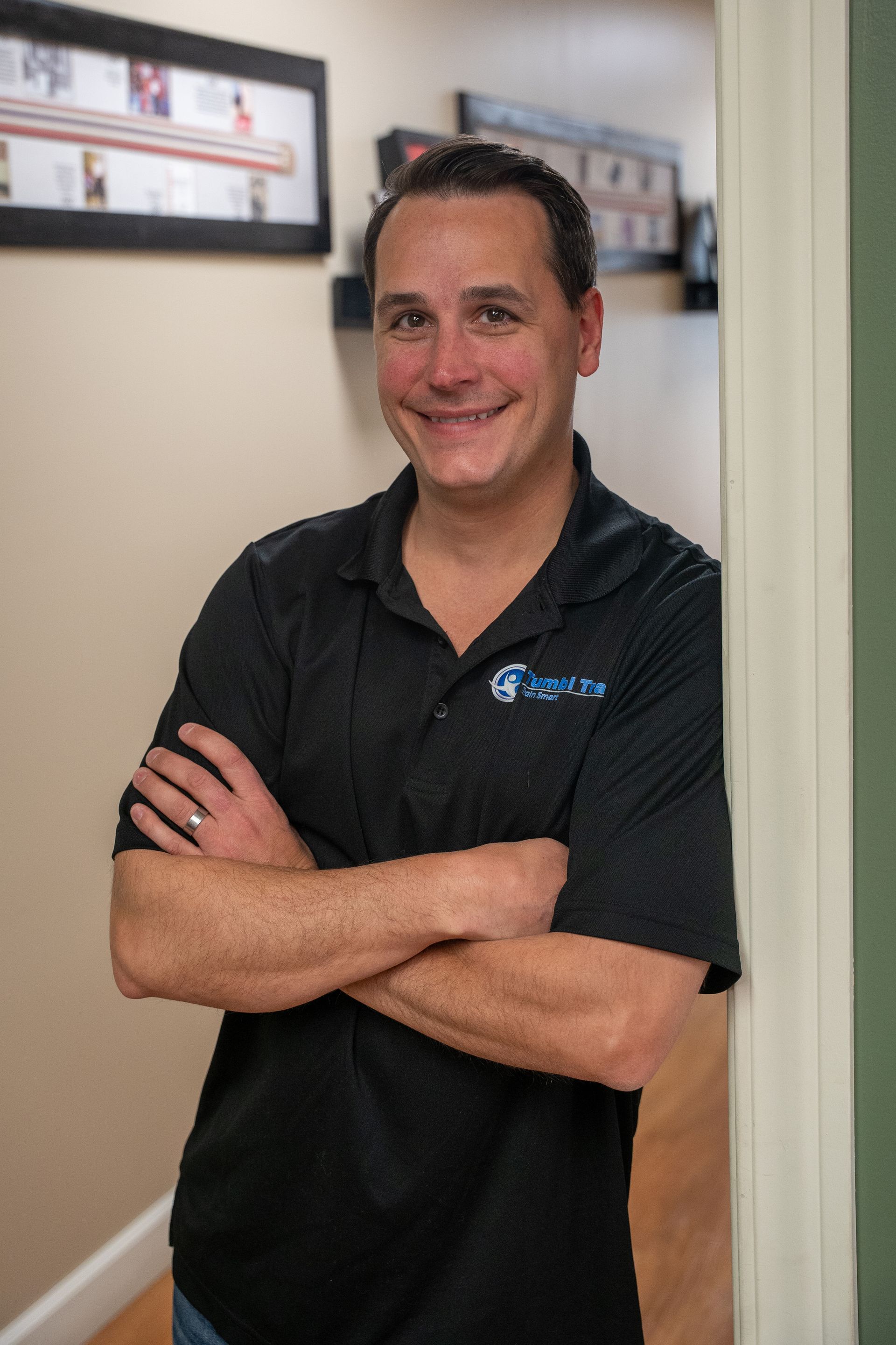 Man standing with arms crossed in a hallway, wearing a black polo shirt and smiling.