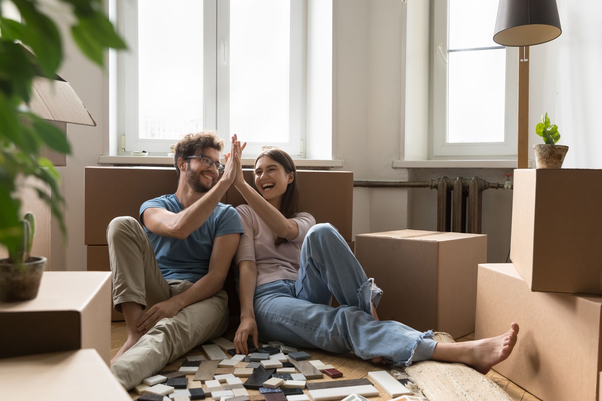 Couple sitting on the floor in a bright room, high-fiving amid moving boxes and scattered packing materials