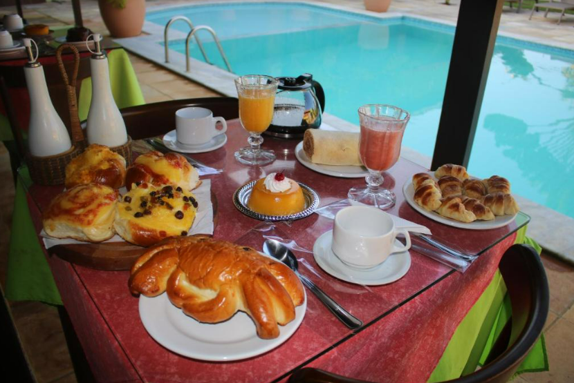 Café da manhã servido à beira da piscina: doces, suco, café e uma pequena sobremesa sobre uma mesa com toalha vermelha.
