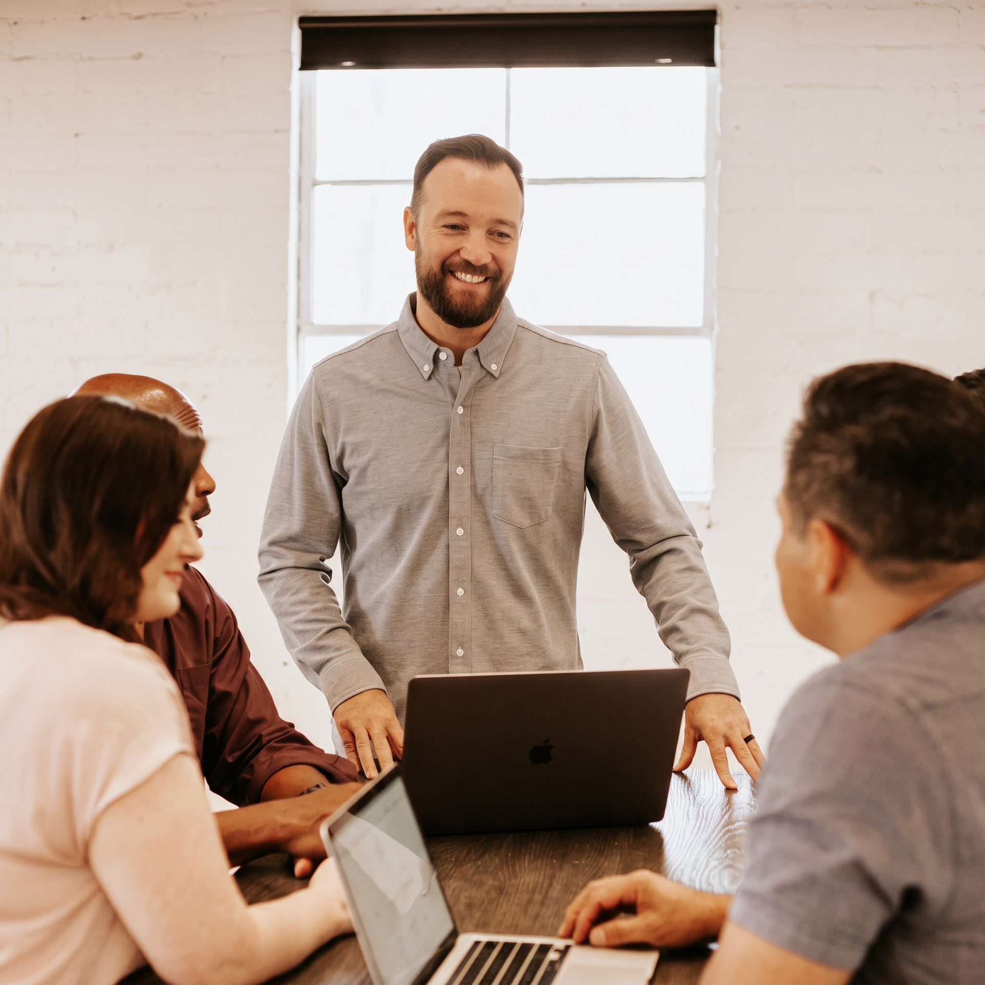 Group meeting around a laptop, with a man standing and smiling in a bright office room