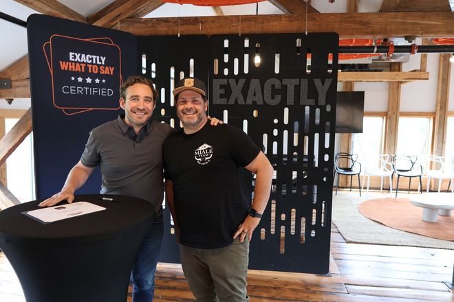 Two men posing at a table in front of an “Exactly” sign at an indoor event booth.