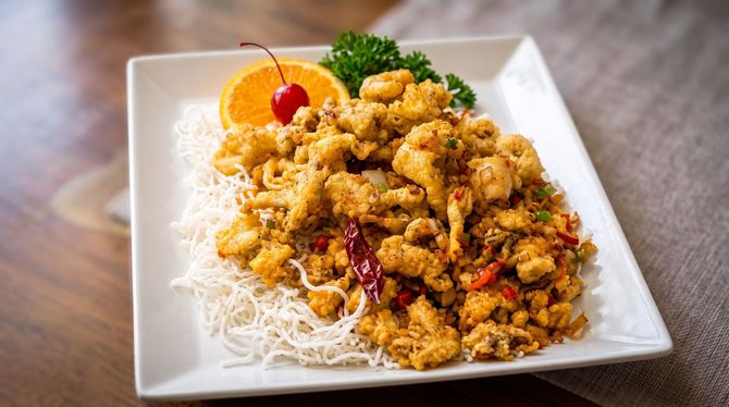 Crispy fried chicken over white noodles on a square plate, garnished with orange slice and parsley