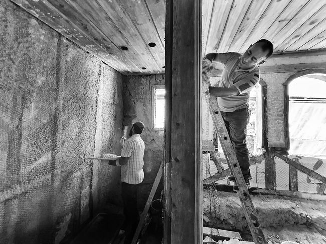 Two workers in a rough, unfinished room, one standing on a ladder by a window and one at the wall.